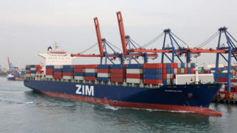 ZIM container ship passing harbor cranes and stacked containers.
