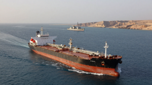 Oil tanker passing through the Strait of Hormuz with a naval vessel in the distance